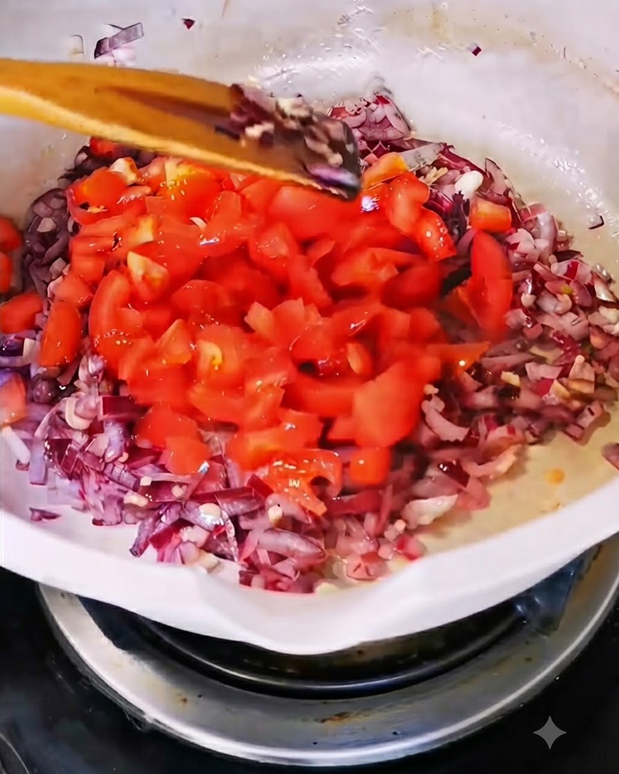 Tomates rojos recién picados añadiéndose en una sartén blanca que contiene cebolla y ajo salteados.
