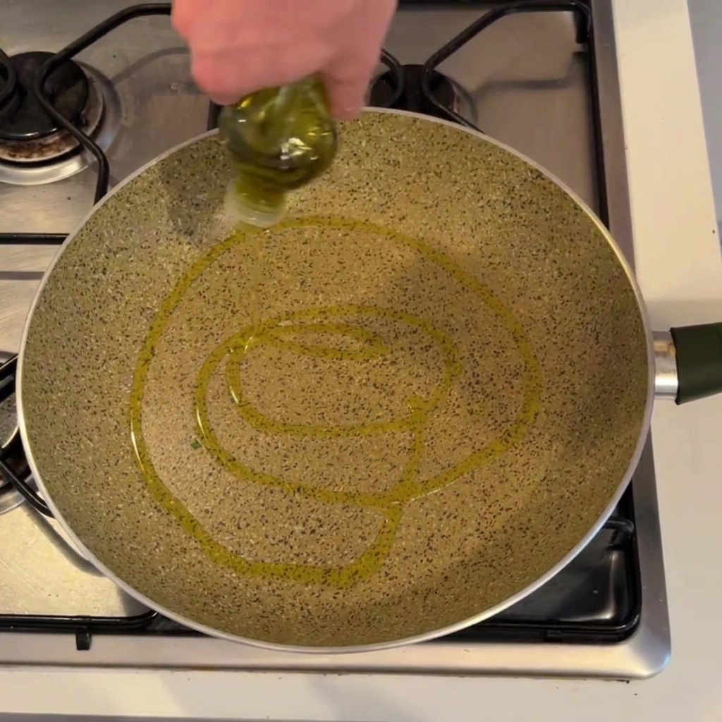 Alguien vertiendo aceite de oliva de una botella en una sartén grande sobre una cocina de gas.