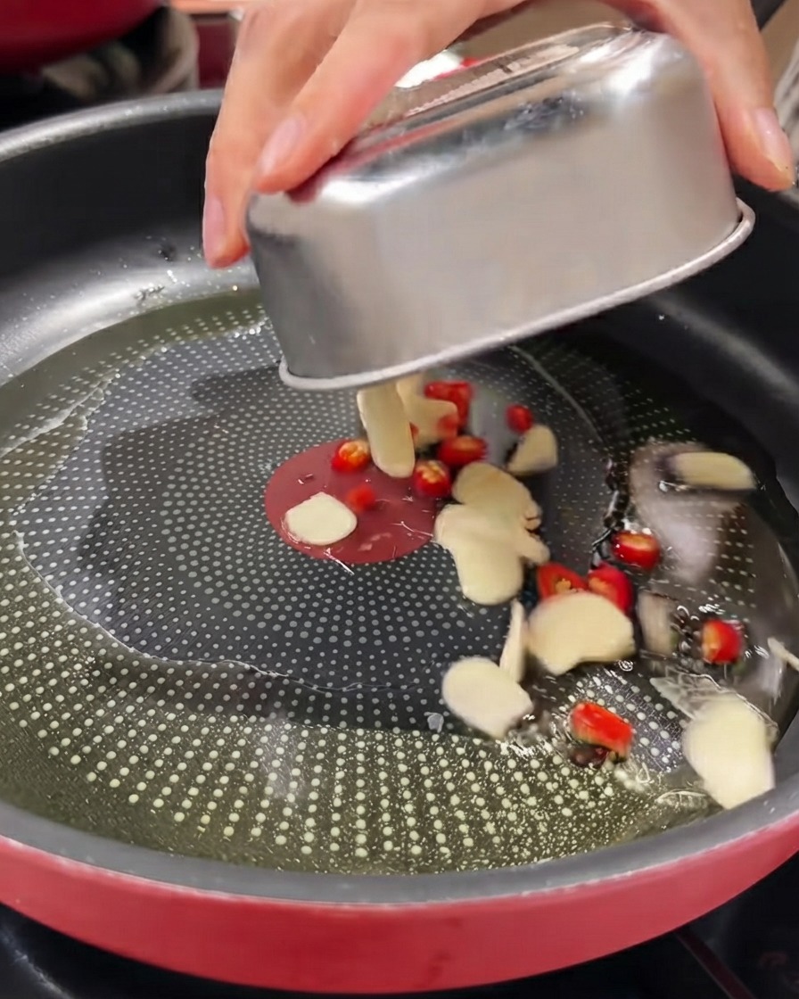 Ajo en rodajas, chiles ojo de pájaro rojos y cebollín siendo vertidos desde un recipiente metálico en una sartén con aceite.
