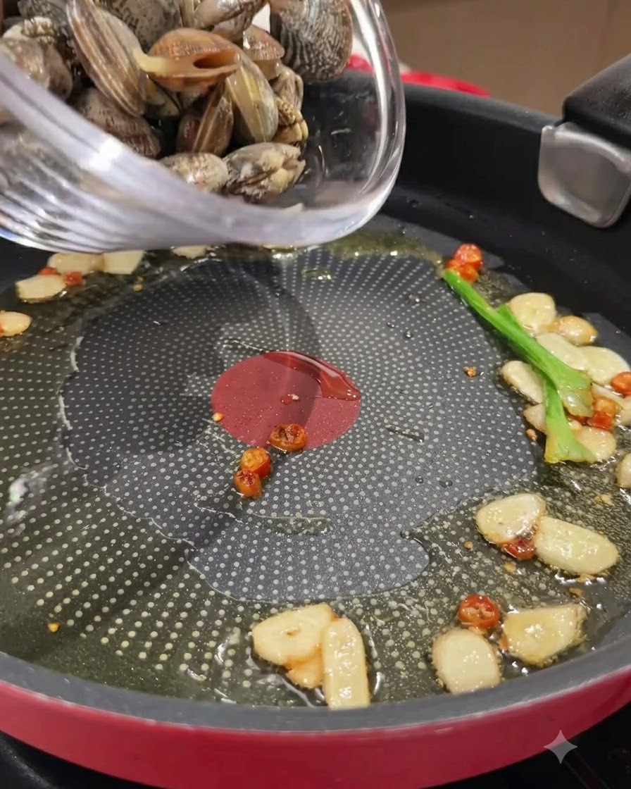 Almejas frescas siendo vertidas desde un tazón de vidrio en una sartén con ajo y chiles salteados en aceite.