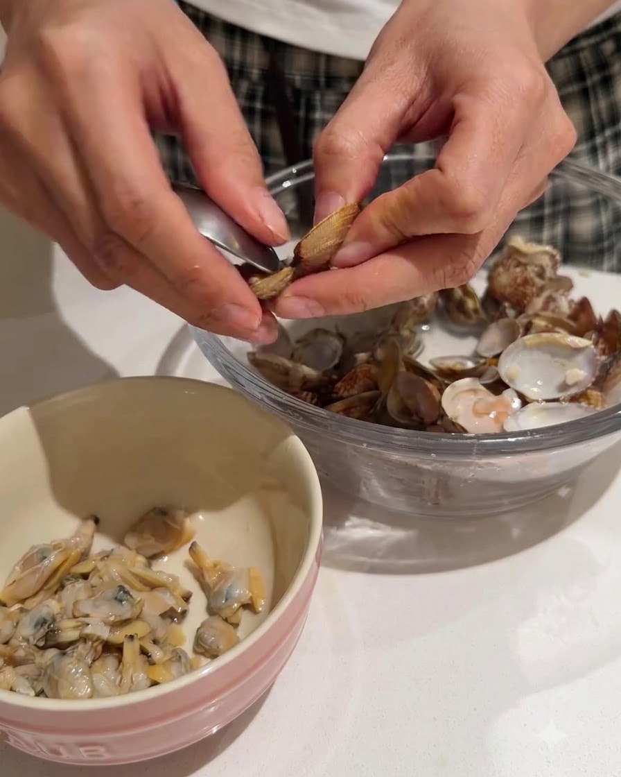 Manos extrayendo la carne tierna de las almejas de sus conchas hacia un bol con borde rosa.
