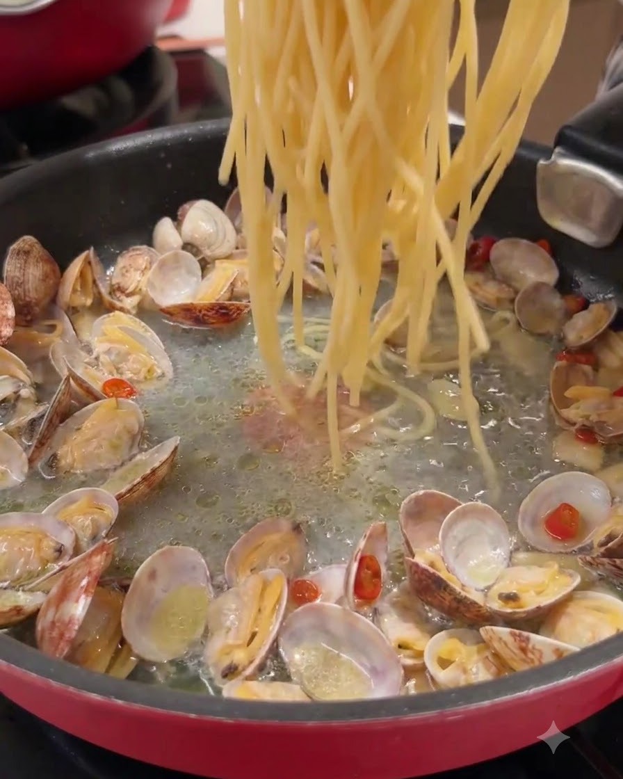 Hebras largas de pasta siendo arrojadas a una sartén que contiene almejas cocidas y líquido sazonado.