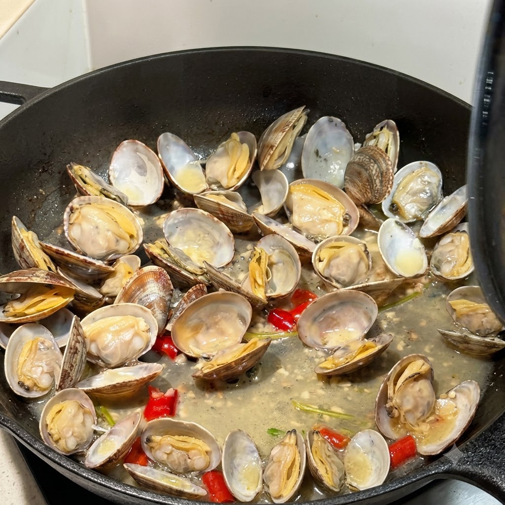 Almejas abiertas cocinándose en caldo dentro de una sartén con una tapa parcialmente visible.
