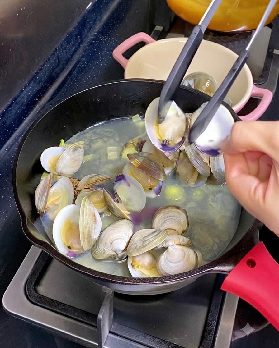 Un par de pinzas de cocina sosteniendo abierta la concha de una almeja con carne cocida dentro, sobre una sartén de almejas y caldo humeantes.