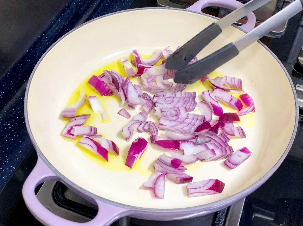 Una sartén de color claro que contiene aceite de oliva y cebollas rojas picadas siendo revueltas con pinzas.