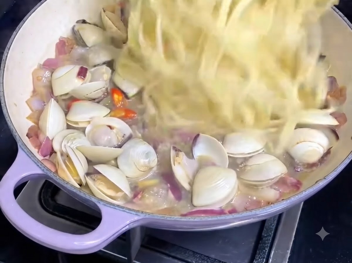 Hebras de espaguetis cocidos siendo incorporadas a una sartén que contiene salsa de almejas y cebollas.