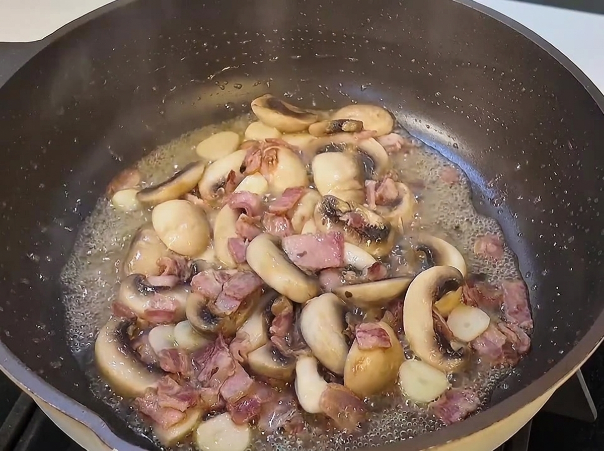 Fideos de espagueti cocidos integrándose en una sartén grande con rodajas de champiñones salteados y trozos de tocino.