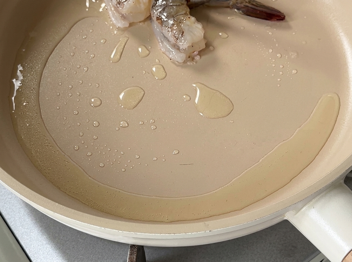 Aceite caliente brillando en una sartén poco profunda con un par de piezas de gamba comenzando a cocinarse.