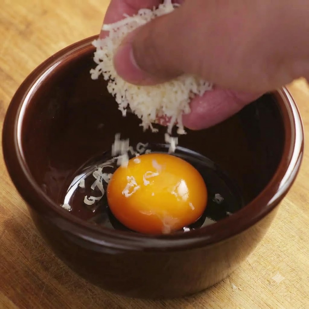 Queso parmesano rallado cayendo sobre una yema de huevo brillante dentro de un tazón de cerámica marrón.