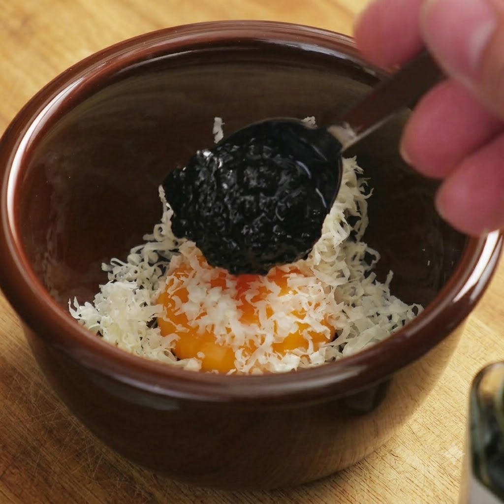 Una cucharada de salsa oscura de trufa negra cayendo sobre la mezcla de yema de huevo y queso.