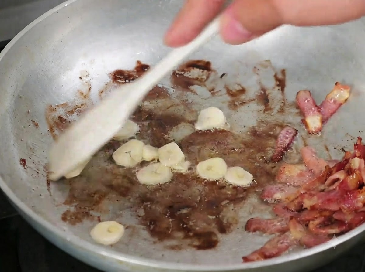 Ajo en láminas mezclándose en la sartén con el tocino cocido usando una cuchara de madera.