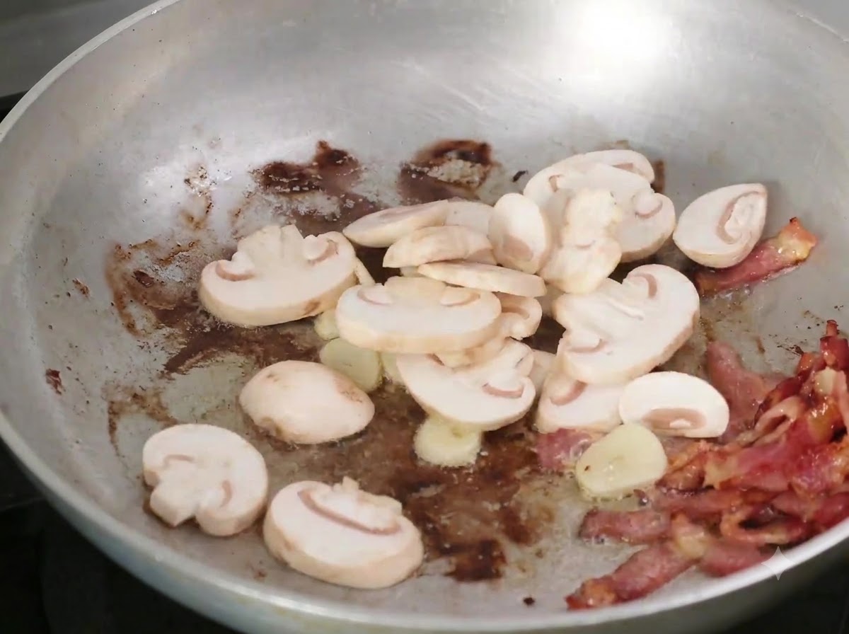 Champiñones en rodajas añadiéndose a la sartén con el tocino y el ajo.