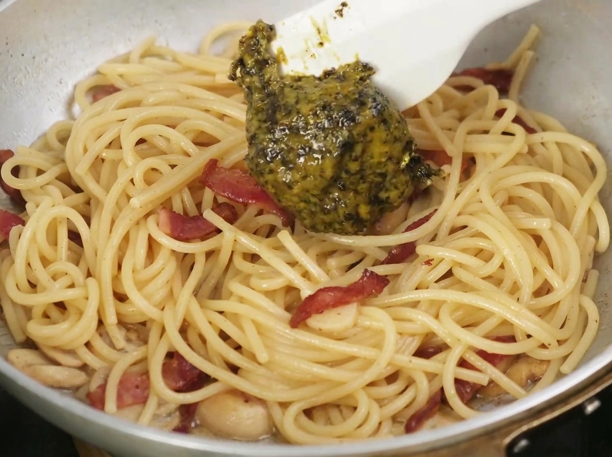 Salsa de trufa negra añadiéndose con una cuchara a una sartén con pasta cocida y tocino.