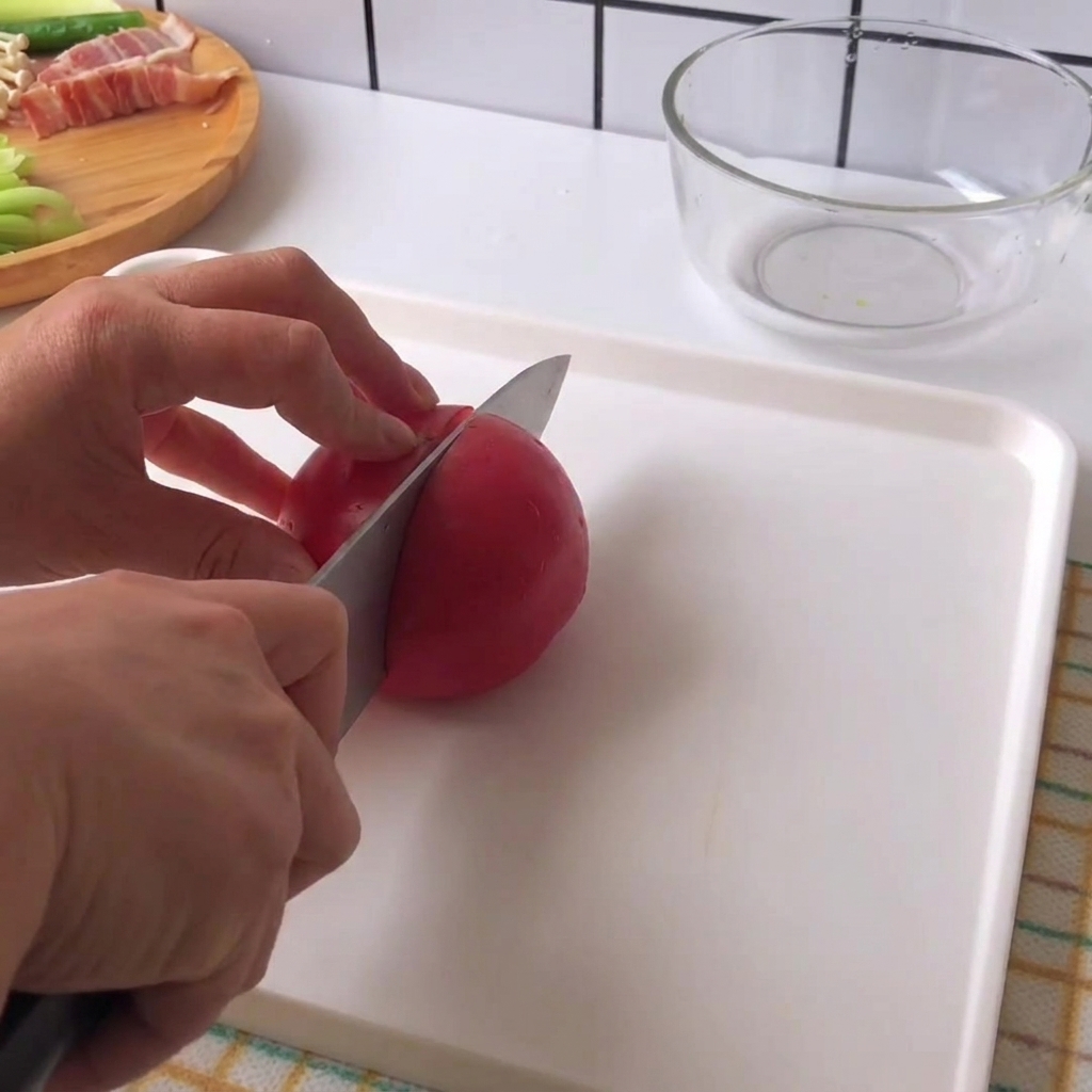 Manos sosteniendo un cuchillo y cortando un tomate rojo fresco en trozos sobre una tabla de cortar blanca.