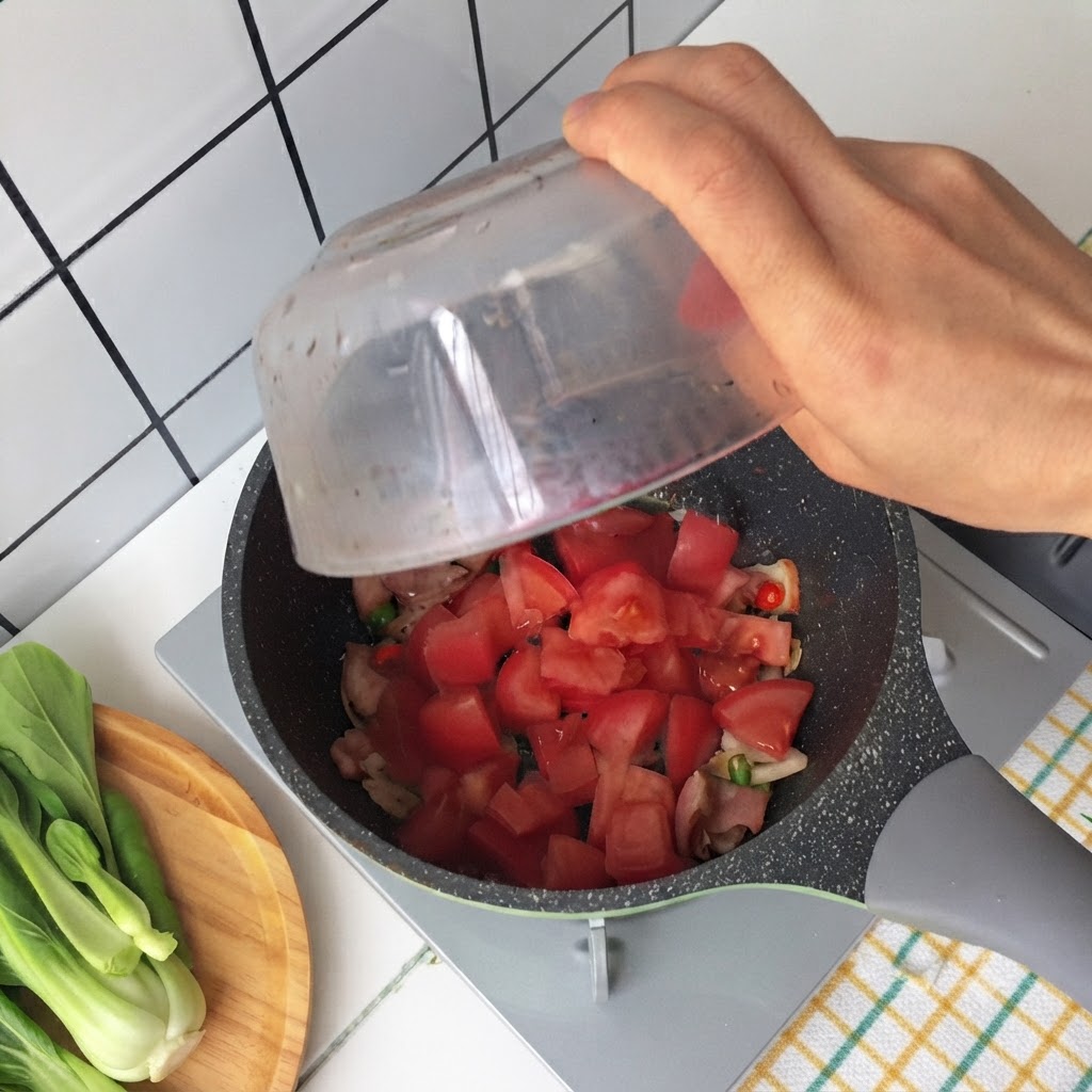 Mano vertiendo tomates picados de un tazón de vidrio a una sartén que contiene tocino frito y cebolletas.