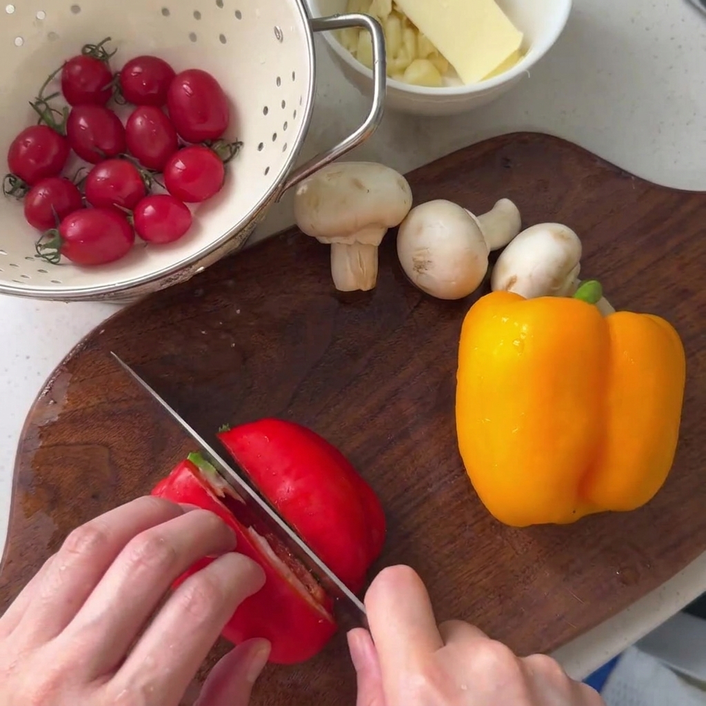 Pimiento rojo cortándose en una tabla de cortar de madera junto a un pimiento amarillo y champiñones botón.