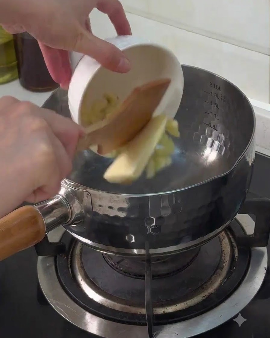 Ajo picado y un trozo de mantequilla vertiéndose desde un tazón blanco pequeño en una olla metálica en la estufa.