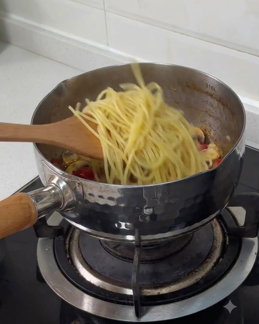 Espaguetis hervidos transfiriéndose con una espátula de madera a una olla de acero inoxidable con otros ingredientes cocinados.