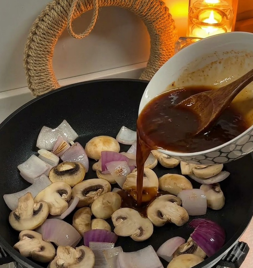 Vertiendo salsa marrón desde un tazón hacia una sartén que contiene cebollas cortadas y champiñones blancos.