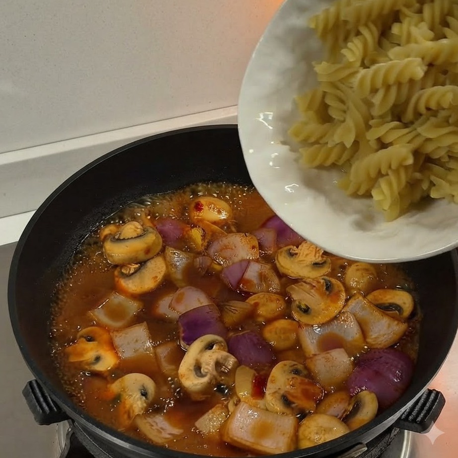 Pasta fusilli hervida siendo vertida desde un plato blanco hacia una sartén llena de salsa y verduras a fuego lento.
