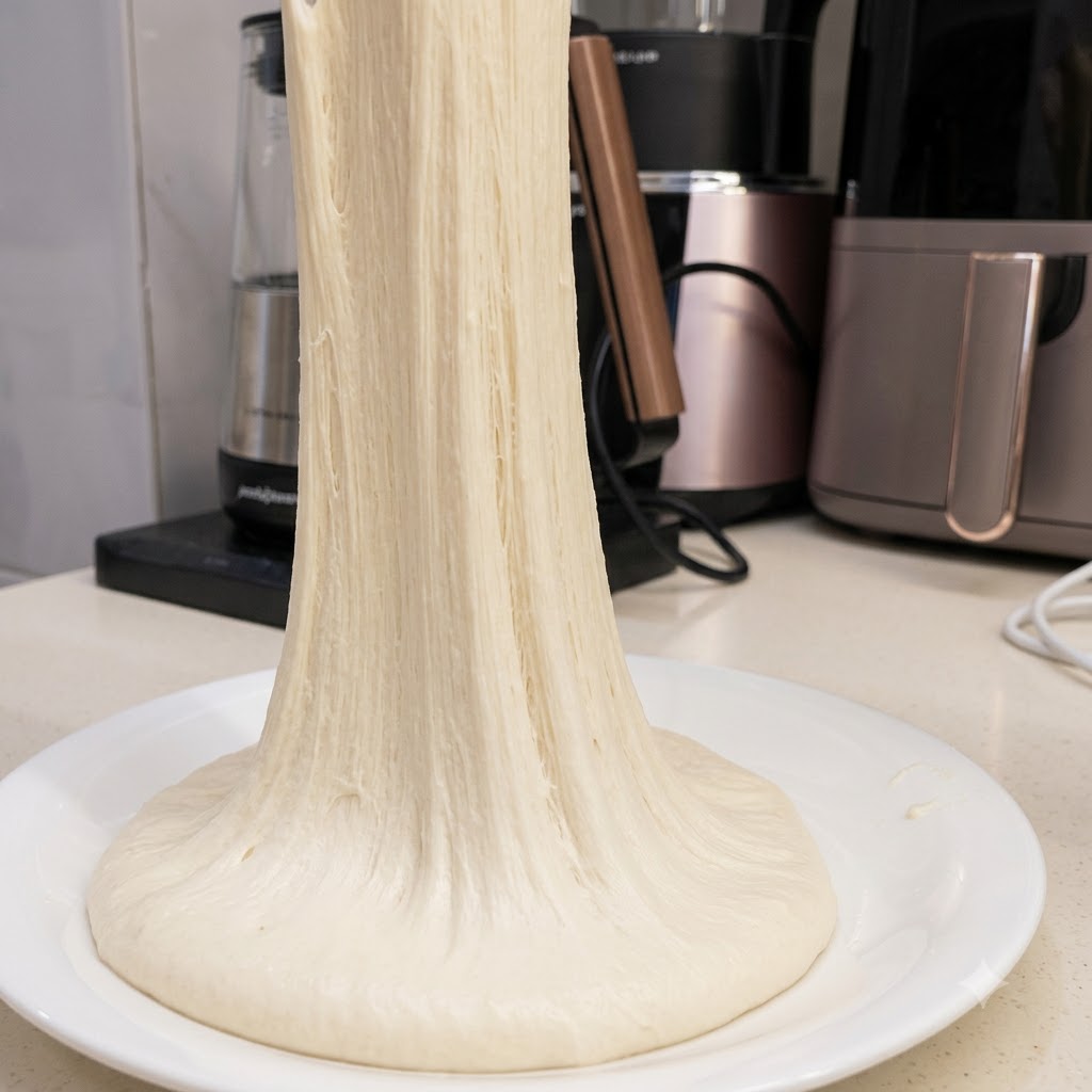 Un filamento largo y continuo de masa de pizza muy elástica que se estira hacia arriba desde un plato blanco.