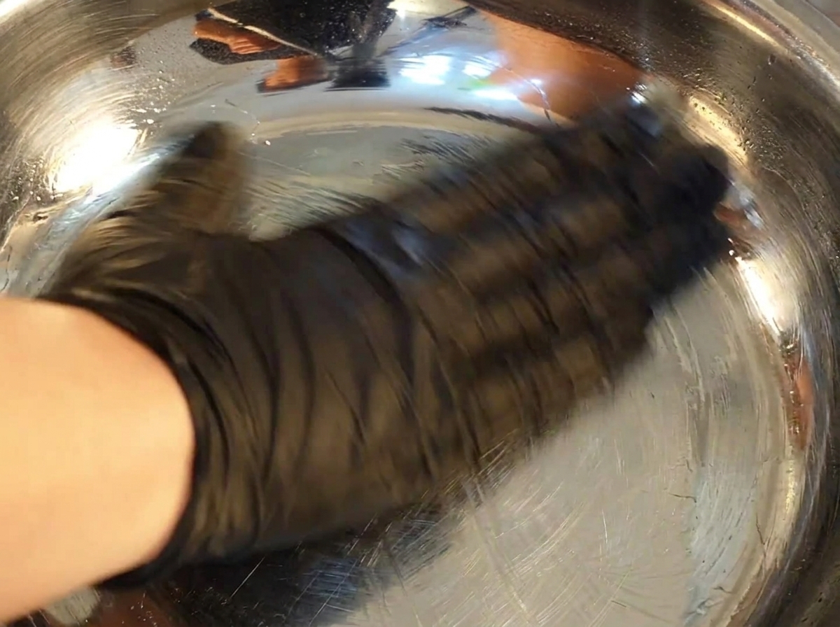 Una mano con un guante negro usa una toalla de papel para cubrir con aceite el interior de un bol de acero inoxidable brillante.