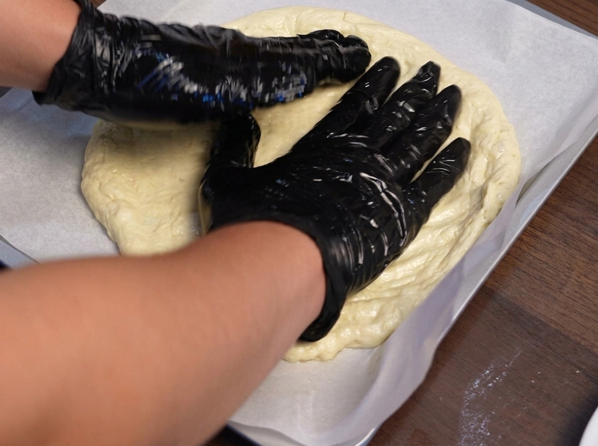 Manos con guantes estirando la masa de pizza hacia afuera sobre una bandeja para hornear forrada con papel vegetal.