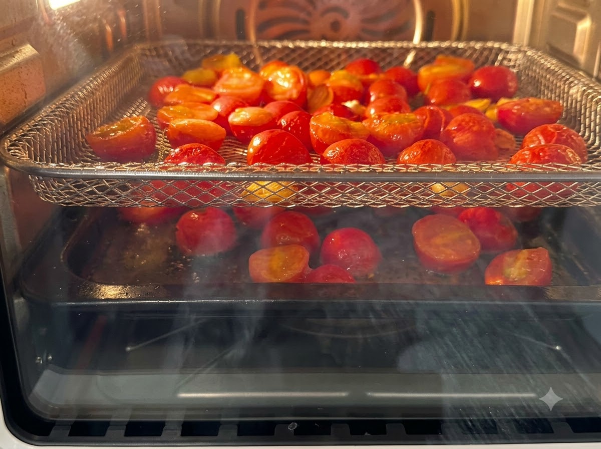 Tomates cherry cortados a la mitad dispuestos en rejillas de freidora de aire horneándose dentro del aparato.