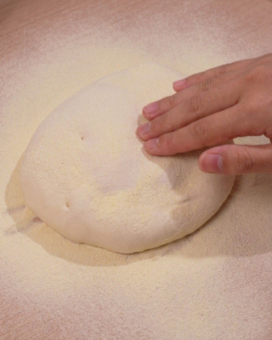 Mano presionando suavemente hacia abajo sobre un montón de masa de pizza que descansa sobre sémola de maíz amarilla.