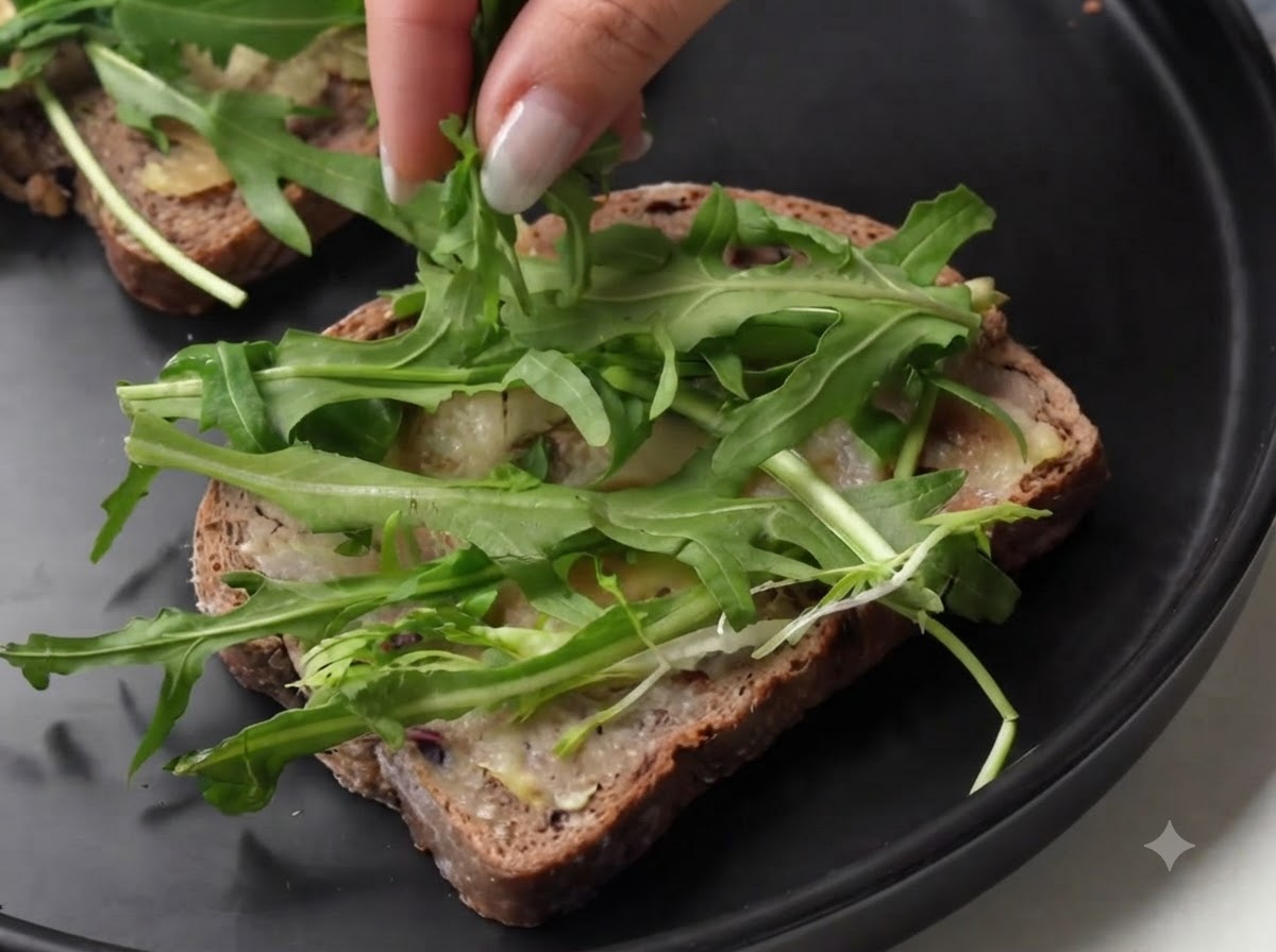 Persona colocando lonchas de jamón curado sobre rúcula verde fresca en una rebanada de pan.