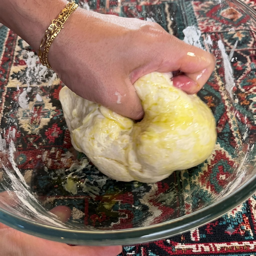Una mano amasando y doblando firmemente masa mezclada con aceite de oliva dentro de un bol de vidrio.
