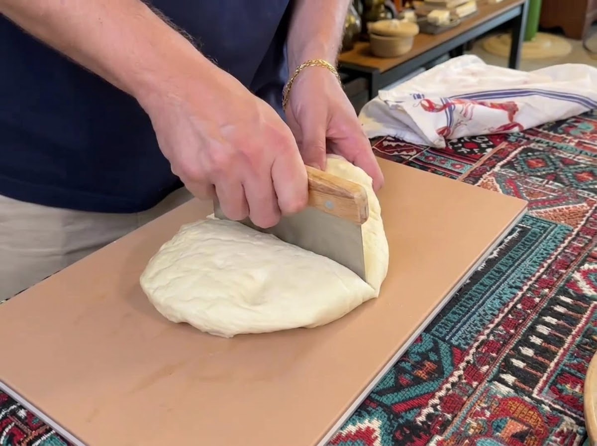Un cornet o rasqueta cortando una gran bola de masa de pizza blanca en cuatro partes separadas sobre una tabla de cortar.