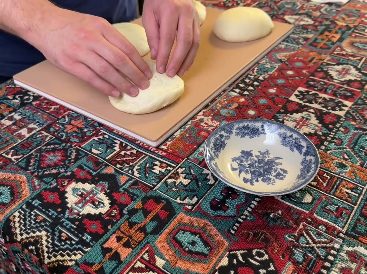 Manos presionando y aplanando una bola redonda de masa de pizza sobre una tabla de madera.