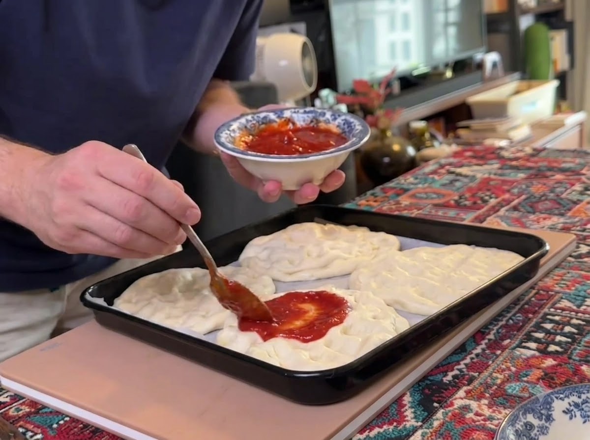Una cuchara extendiendo salsa de tomate roja sobre múltiples bases de masa de pizza aplanadas dispuestas en una bandeja de horno negra.