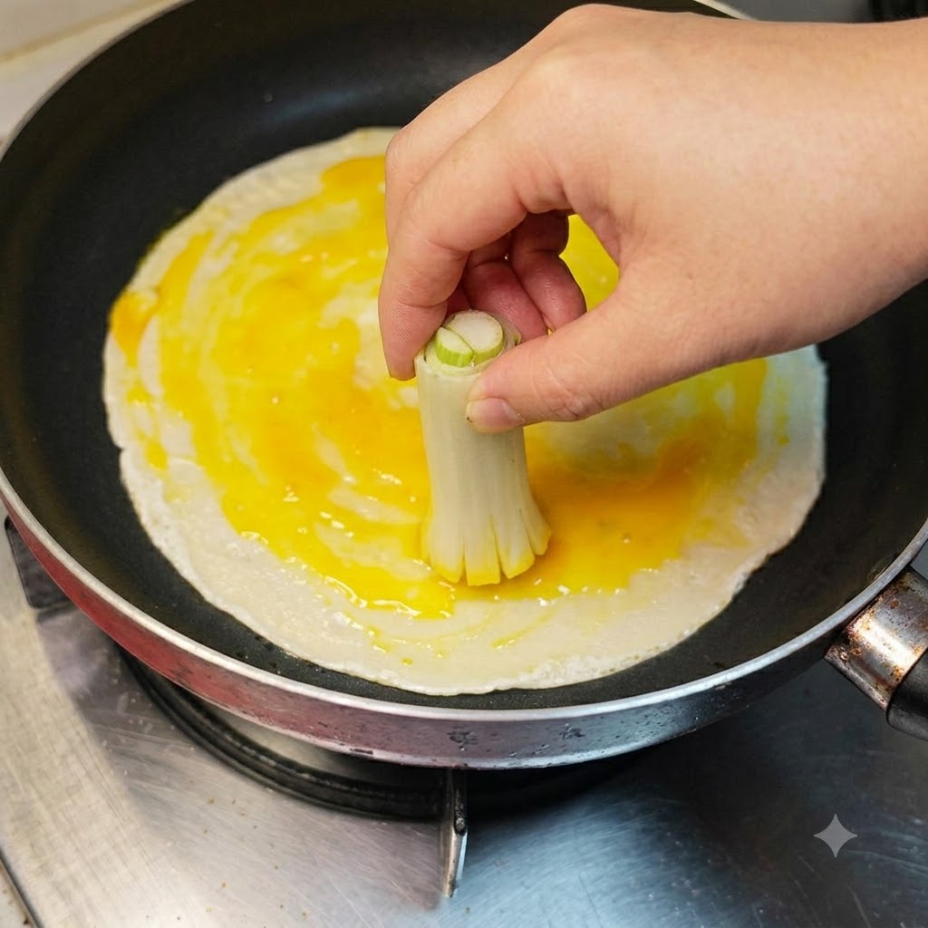 Una mano usando un manojo atado de tallos de cebollino para esparcir la clara y yema de huevo batida a través de un panqueque.