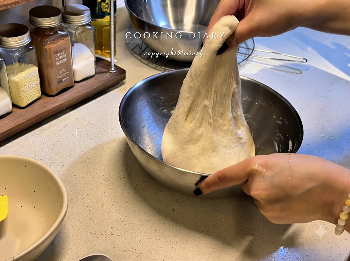 Un par de manos levantando una porción de masa de pizza elástica hacia arriba dentro de un bol de metal para estirarla.
