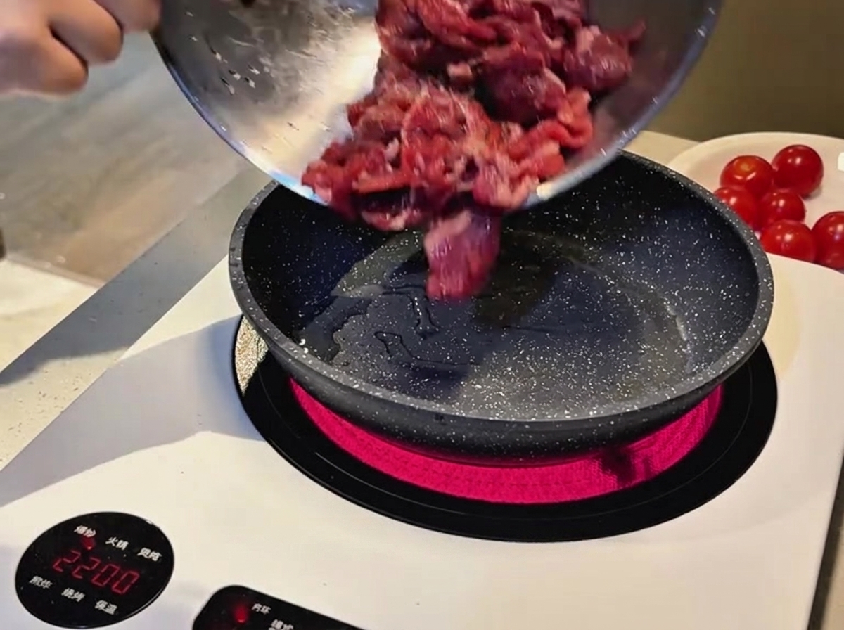 Tiras de ternera cruda marinada cayendo de un bol de metal a una sartén caliente en una placa de inducción.