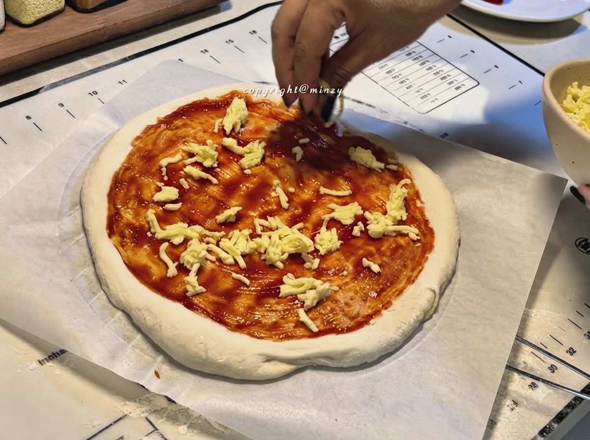 Manos espolvoreando queso rallado sobre una base de pizza cubierta con salsa roja.