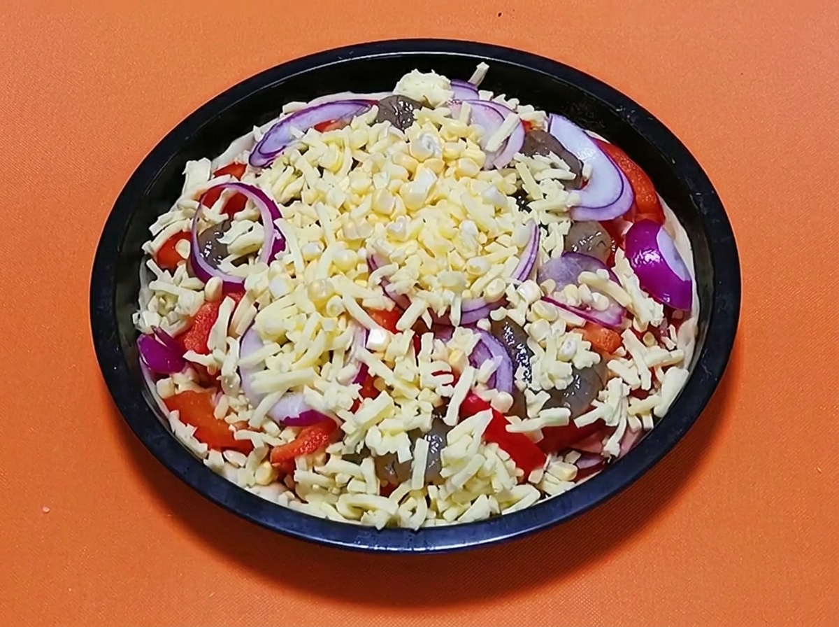 Pizza de mariscos cruda completamente armada cubierta con una capa final de queso rallado y rodajas de cebolla roja, lista para hornear.