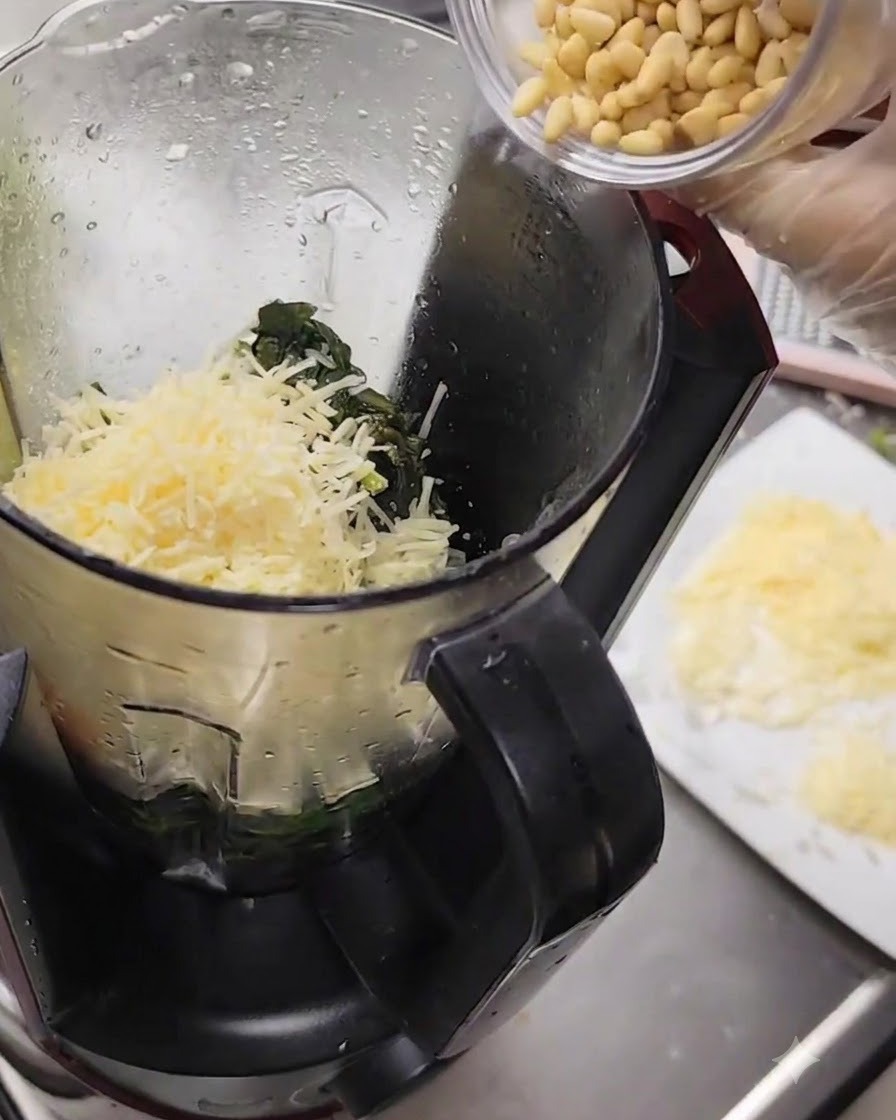 Piñones vertidos desde un vaso pequeño a la licuadora llena de queso y albahaca.
