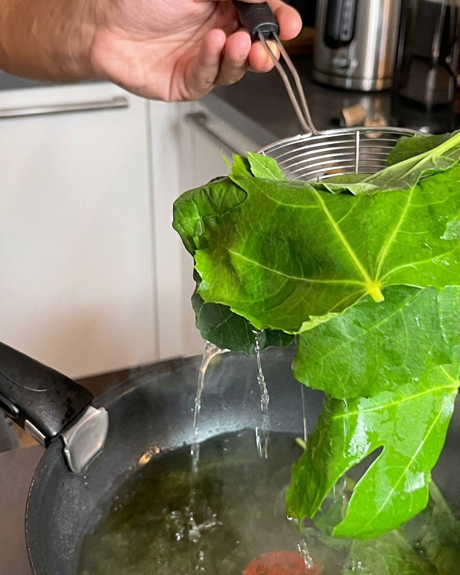 Un colador levantando hojas verdes recién blanqueadas de una olla con agua hirviendo.