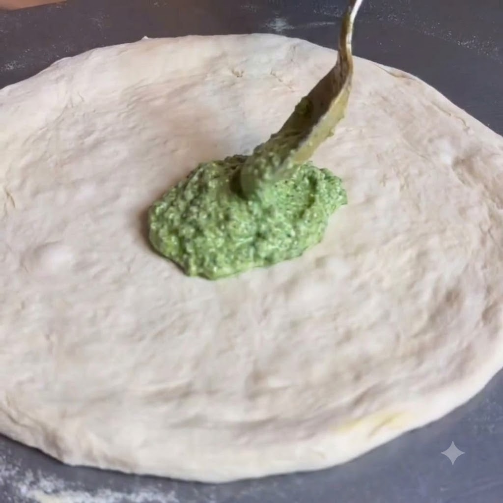 Una cuchara extendiendo salsa de pesto verde por el centro de una masa de pizza plana y redonda.