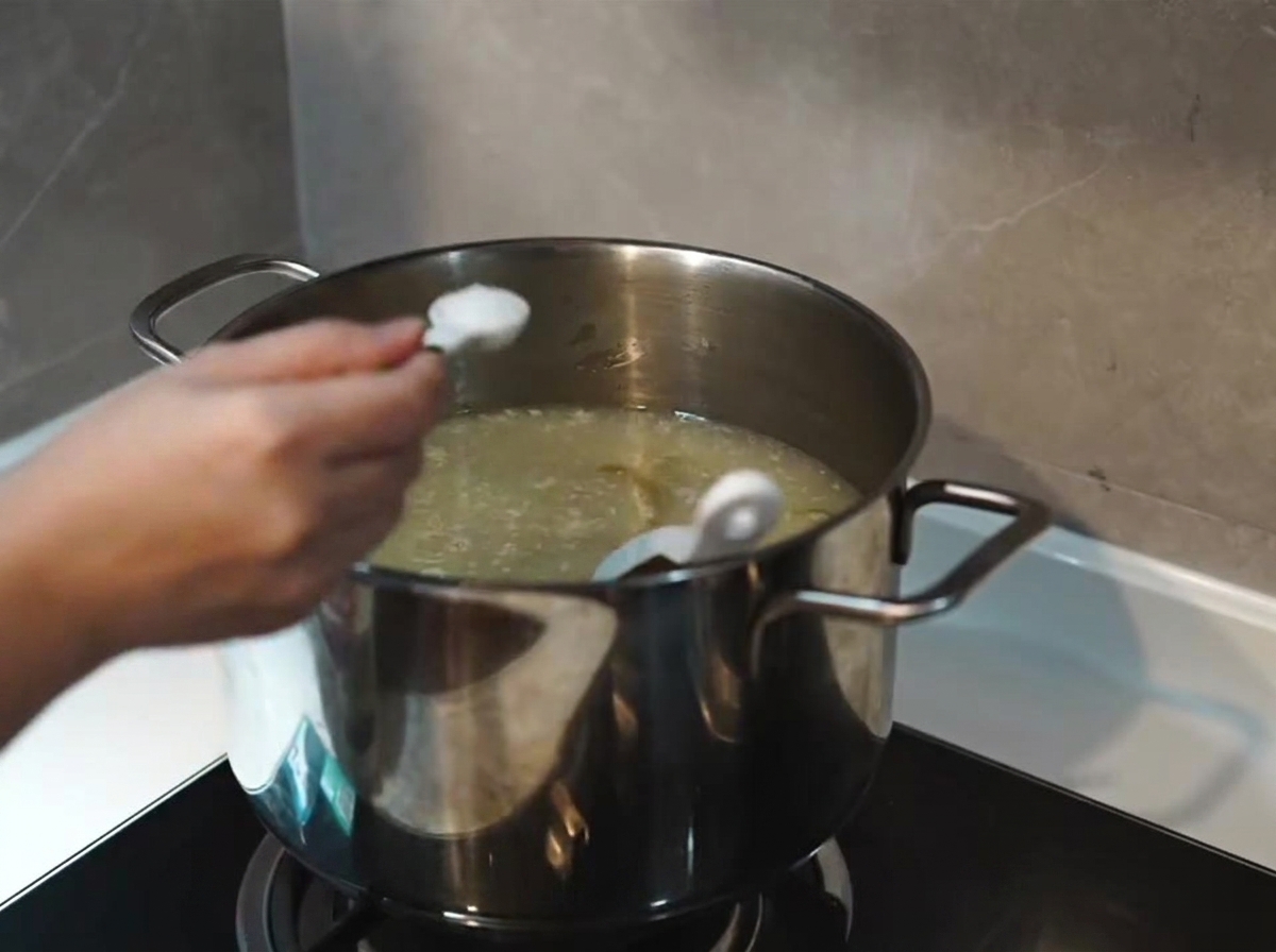 Una cucharada de sal siendo añadida a una base de sopa blanca lechosa, espesa y hirviendo en una olla de acero inoxidable.