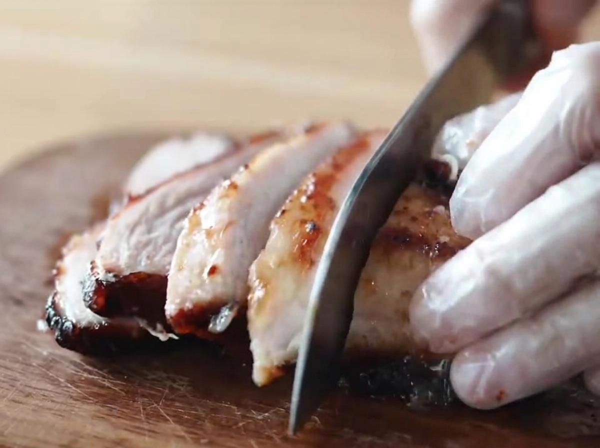 Un trozo de cerdo chashu ahumado caramelizado y glaseado con miel siendo cortado en trozos gruesos por alguien con guantes sobre una tabla de cortar de madera.