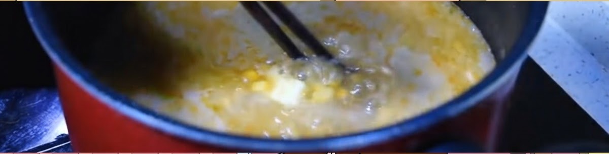 Palillos removiendo un caldo de miso naranja hirviendo vigorosamente con maíz y una porción de mantequilla en una olla oscura.