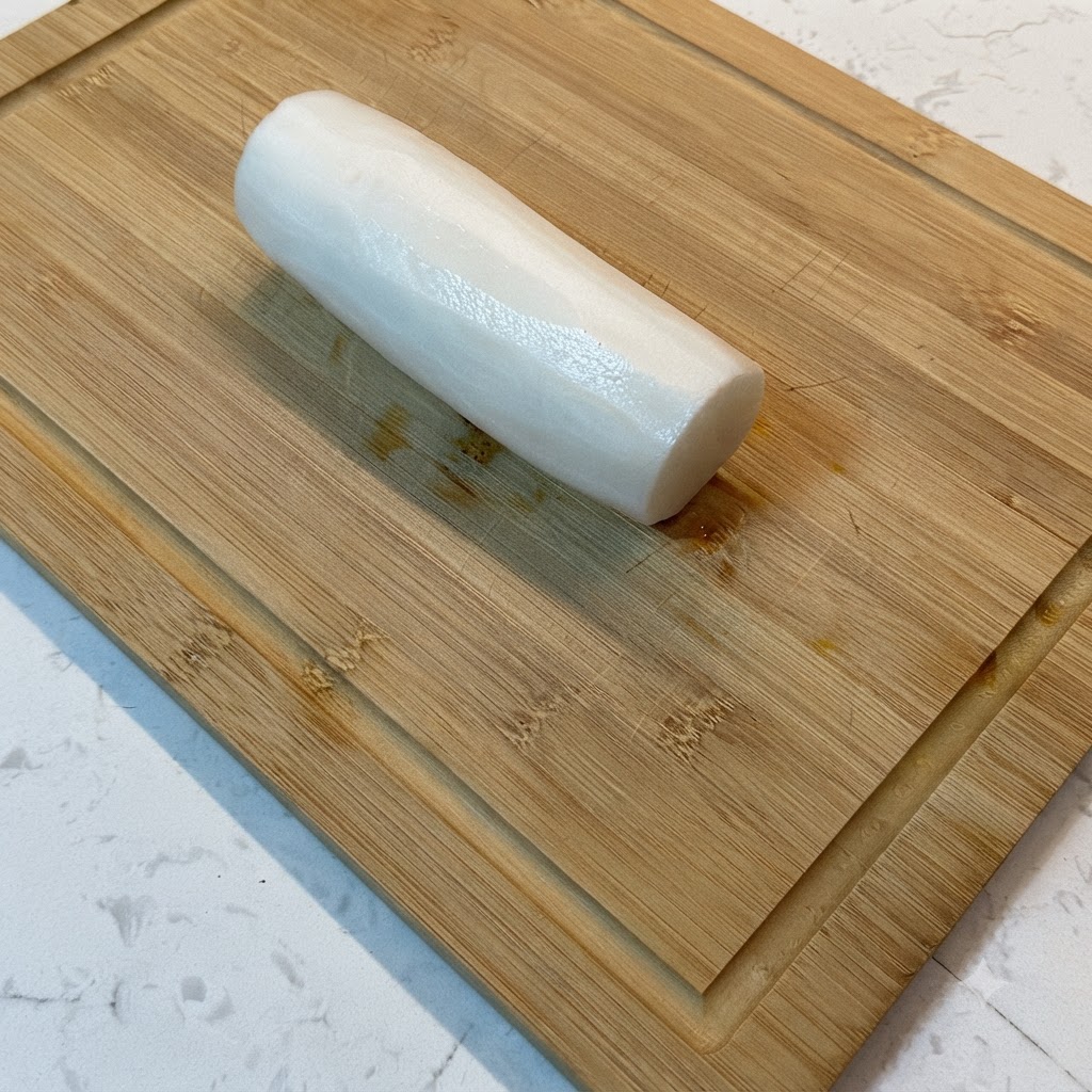 Un rábano blanco siendo cortado en tiras finas sobre una tabla de cortar de madera.