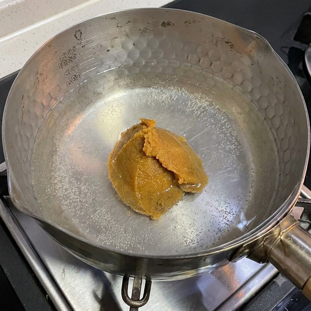 Una cucharada de pasta de miso blanco disolviéndose en una olla con agua caliente en una estufa.
