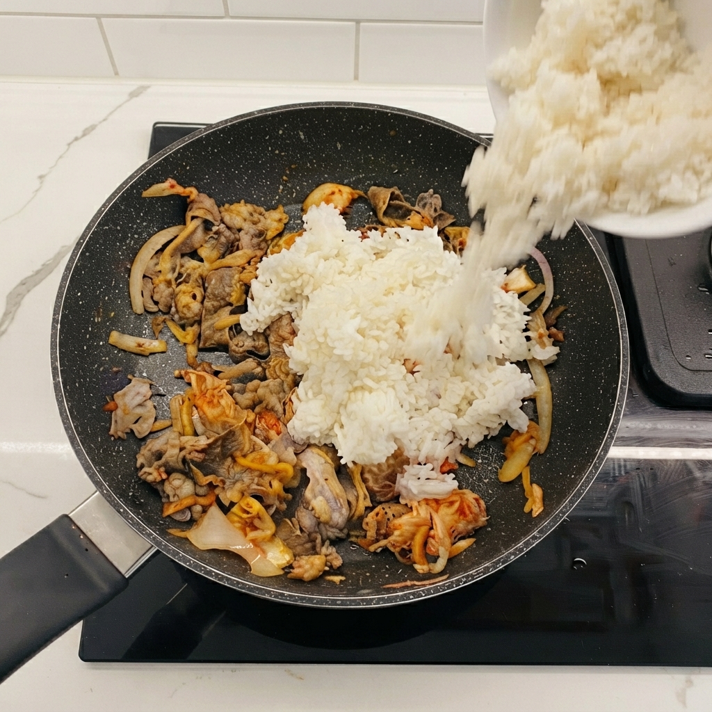 Arroz blanco siendo vertido desde un bol hacia la sartén que contiene ternera y kimchi.