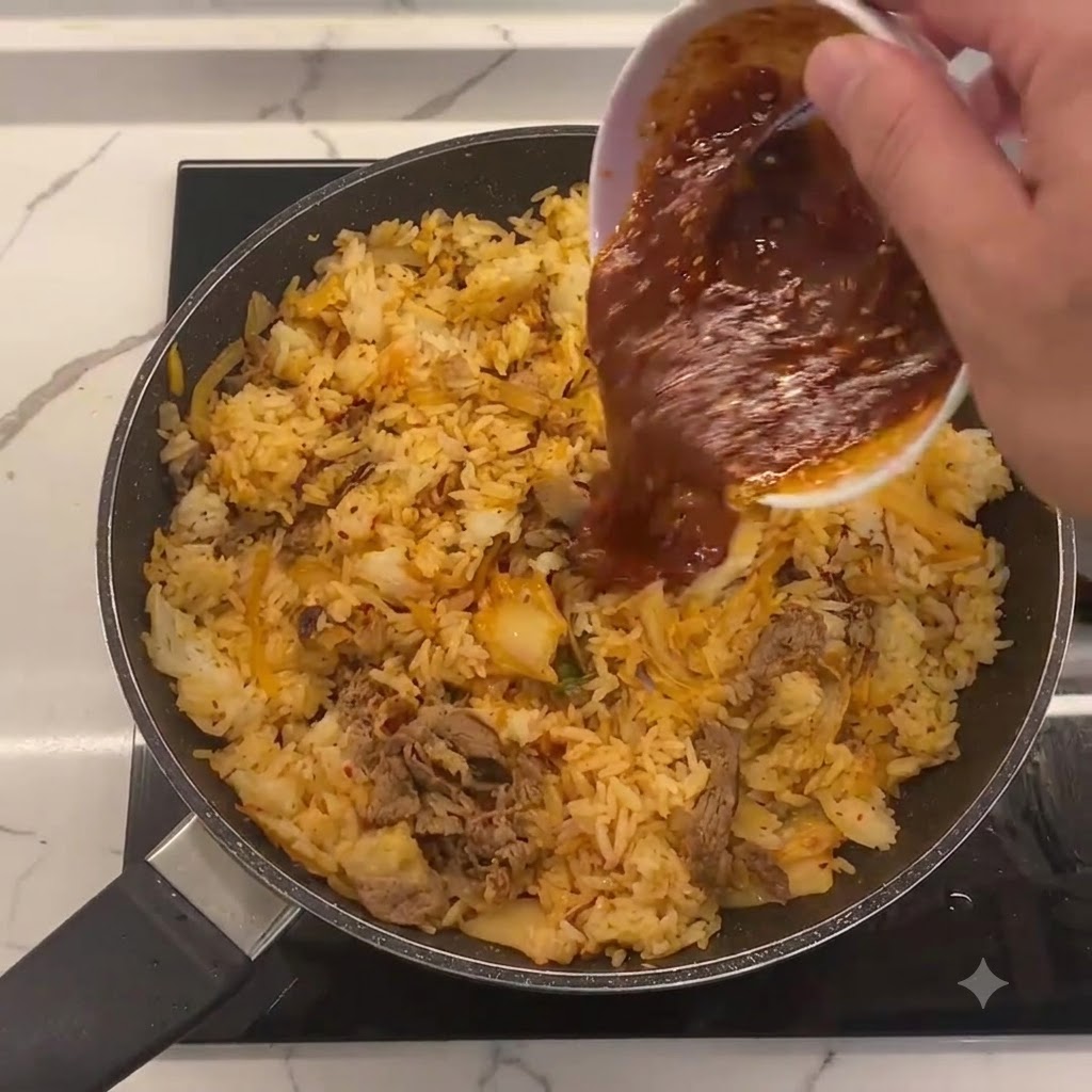 Salsa especial siendo vertida desde un bol blanco pequeño en el centro de la sartén con el arroz frito, el kimchi y la ternera.