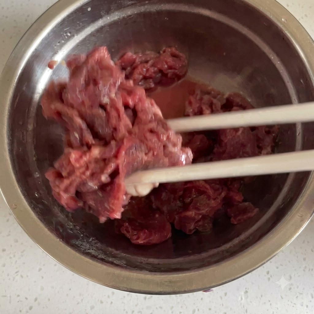 Ternera cruda cortada en rodajas finas mezclándose minuciosamente con los ingredientes del adobo usando palillos dentro de un tazón de acero inoxidable.